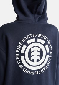 Tmavomodrá mikina s kapucňou s veľkým bielym kruhovým obrázkom na chrbte s elementárnymi symbolmi a motívom stromu v strede. Mäkká textúra.