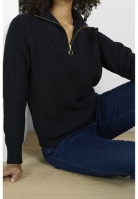 Schwarzer Strickpullover mit hohem Kragen, goldfarbener Reißverschluss und gerippten Ärmelbündchen. Kombiniert mit blauen Jeans.