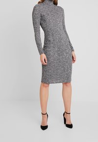 Vaaleanharmaa neulottu mekko pitkillä hihoilla, korkealla kaula-aukolla, vartalonmyötäisellä siluetilla, polvenpituinen, sileä pinta, yhdistetty mustiin korkeakorkoisiin kenkiin.