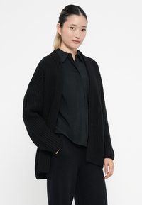 Svart stickad cardigan med ribbad struktur, öppen front och sänkta axlar, bärs över en mörk, knäppt skjorta. Mjuk, avslappnad passform.