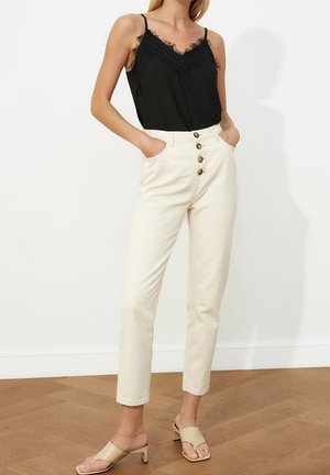 Vrouw draagt een zwart kanten hemdje, ingestopt in een hooggerichte, off-white broek met knopen aan de voorkant, staand op een houten vloer.