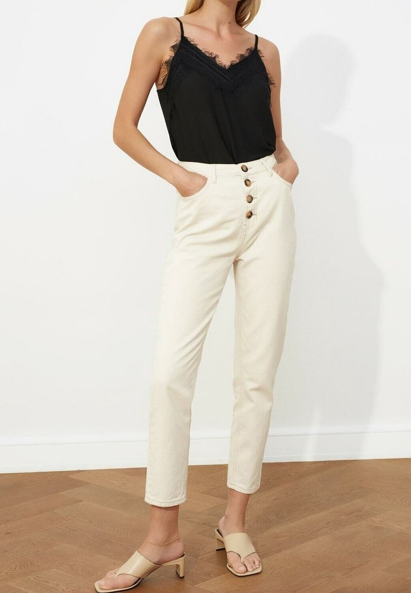 Femme portant une camisole noire à bordure en dentelle rentrée dans un pantalon taille haute écru avec des boutons à l'avant, debout sur un sol en bois.