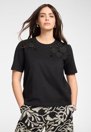 Kobieta z długimi ciemnymi włosami nosi czarną koszulkę z krótkim rękawem ozdobioną koralikowymi kwiatowymi wzorami na ramionach oraz czarno-białe spodnie w wzory.