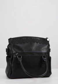Sac à main en cuir noir avec bord festonné, motif floral embossé, poche avant zippée et doubles poignées sur fond gris.