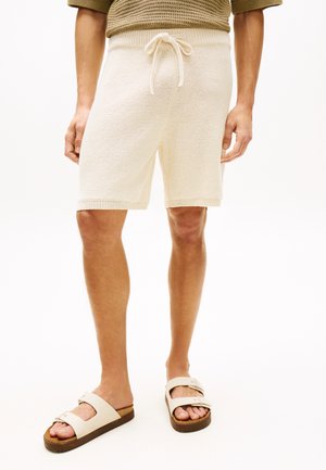Mann trägt cremefarbene Strickshorts mit Kordelzug und beige Slides, steht vor weißem Hintergrund.