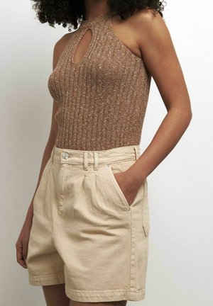 Vrouw draagt een mouwloos bruin geribbeld topje met een keyhole halslijn en beige shorts met hoge taille, één hand in de zak.