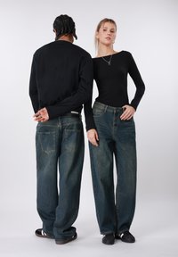 Μαύρο μακρυμάνικο τοπ και ξεβαμμένο μπλε φαρδύ τζιν σε δύο άτομα, που προβάλλουν μια χαλαρή εφαρμογή και ιδιαίτερες λεπτομέρειες στις πίσω τσέπες.