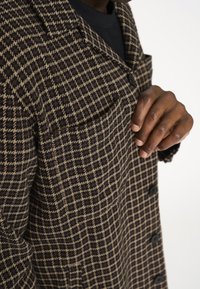 Knapfrakke med hundetandsmønster i brun og beige, lavet af tekstureret stof med en enkelt brystlomme og sorte knapper.