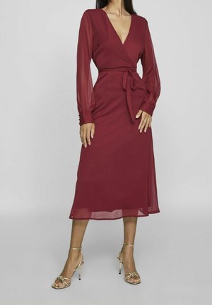 Woman wearing a long burgundy wrap dress with sheer long sleeves and gold strappy high-heeled sandals standing against a gray background.
