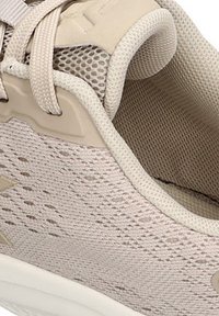 Zapatilla deportiva de color beige con parte superior de malla, patrón texturizado, acentos suaves y cordones blandos. La suela es blanca con un diseño sutil.