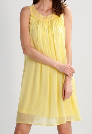 Vrouw in een mouwloze gele jurk tot op de knie met geplooide stof en een gestructureerde halslijn, staand met de armen licht gekruist.