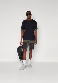 Άνδρας που φοράει μαύρα υπερμεγέθη γυαλιά ηλίου, μαύρο μπλουζάκι, πράσινο και μαύρο σορτς, λευκές κάλτσες, λευκά αθλητικά παπούτσια, κρατώντας μαύρο σακίδιο.