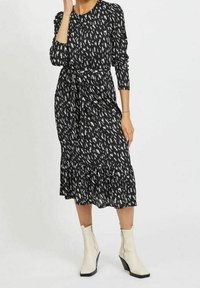 Musta midi-mekko valkoisella abstraktilla kuviolla, jossa on pitkät hihat, sidontavyö ja kerrostettu röyhelöhelma. Yhdistetty vaaleanbeigeihin nilkkureihin.