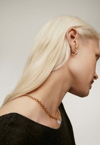Collar de cadena dorada con un colgante ovalado, combinado con un arete de aro dorado retorcido, todo sobre una blusa negra sin hombros.