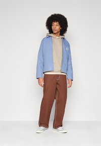 Giacca azzurro chiaro sopra un felpa beige, abbinata a pantaloni marroni e sneakers bianche. La giacca presenta un logo visibile e una vestibilità comoda.