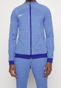 Blå atletisk jakke med lynlås foran, der har tekstureret stof, lilla detaljer ved kraven og manchetterne samt en hvid logo på brystet.