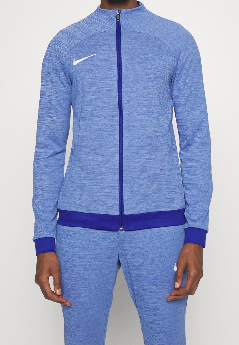 Blå atletisk jakke med lynlås foran, der har tekstureret stof, lilla detaljer ved kraven og manchetterne samt en hvid logo på brystet.