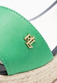 Grön lädersandal med en texturerad yta, som har en logotyp i guldton och ett repdetalj längs basen. Slät foder och kontrasterande sula.