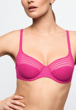 Vrouw die een roze, halftransparante gestreepte bh met dunne bandjes draagt, getoond van schouders tot middel tegen een effen achtergrond.