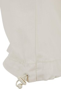 Beige stoffveske med snor-lukking, med en jevn tekstur, sydd sømmer og rund bunn.