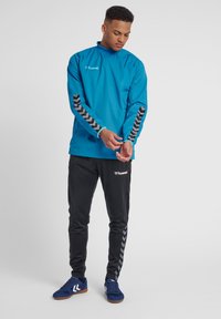 Blauwe windjack met een hoge kraag en zwarte chevronstrepen op de mouwen, gecombineerd met zwarte broek met een logo en bijpassende chevronaccenten.