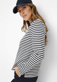 Langärmeliges gestreiftes Shirt in Marineblau und Weiß mit Rundhalsausschnitt, Seitentaschen und lockerer Passform. Kombiniert mit einer marineblauen Mütze.