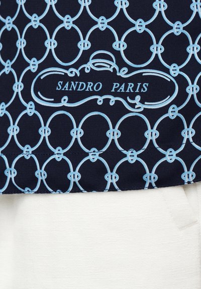 Temno modra tkanina s svetlo modrim prepletenim ovalnim vzorcem in napisom "SANDRO PARIS" nad belo teksturirano tkanino.