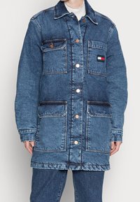 Long denim coat, light blue, with a pointed collar, front buttons, four pockets, and a logo patch on the chest. Distressed texture and seams.