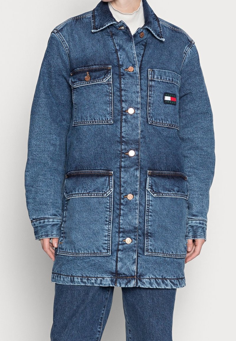 Long denim coat, light blue, with a pointed collar, front buttons, four pockets, and a logo patch on the chest. Distressed texture and seams.