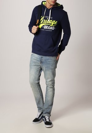 Felpa blu navy con dettagli verdi e testo grafico, abbinata a jeans blu chiaro e sneakers nere. Presenta una tasca frontale e cappuccio con cordino.