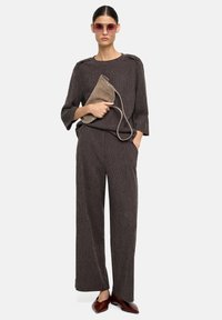 Brązowy sweter w paski z dzianiny z rękawami do połowy łokcia, zestawiony z szerokimi spodniami. Modelka trzyma szarą zamszową torebkę i nosi różowe okulary przeciwsłoneczne.