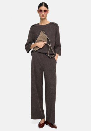 Brązowy sweter w paski z dzianiny z rękawami do połowy łokcia, zestawiony z szerokimi spodniami. Modelka trzyma szarą zamszową torebkę i nosi różowe okulary przeciwsłoneczne.