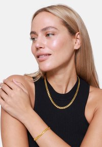 Collier en chaîne dorée et bracelet assorti, avec un design épais et composé de maillons. Porté sur un haut à bretelles côtelé noir. Texture lisse et brillante.