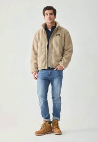 Beige fleecejacka med hög krage, dragkedja fram, kombinerad med blå jeans och bruna stövlar med svarta detaljer.