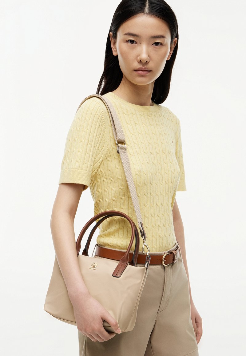 Young woman with long black hair wears yellow knit short-sleeve top, beige pants, and holds beige handbag with brown handles and strap.