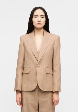 Vrouw met lang donker haar draagt een beige pinstripe blazer met gewatteerde schouders en bijpassende hoge taillebroek tegen een eenvoudige grijze achtergrond.