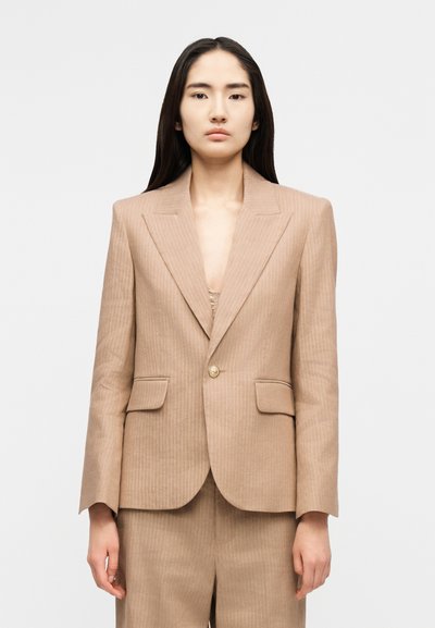 Femme aux longs cheveux noirs portant un blazer beige à rayures fines avec des épaules rembourrées et un pantalon taille haute assorti, sur un fond gris uni.