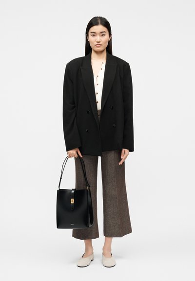 Γυναίκα που φοράει καφέ παντελόνι με ευρεία μπατζάκια, λευκό πουκάμισο με κουμπιά, μαύρο υπερμεγέθη δίπλα φυλλαδωτό σακάκι, κρατώντας μαύρη δερμάτινη τσάντα χειρός με χερούλι στο πάνω μέρος.