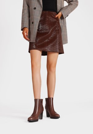 Gonna in pelle marrone con una finitura lucida e texture, abbinata a stivali neri alla caviglia. La modella indossa un blazer a scacchi e un top nero.