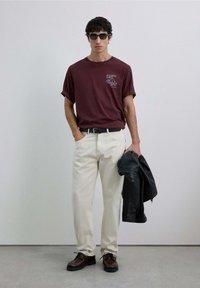 Camiseta burdeos con diseño gráfico, pantalones color crema, zapatos marrones y chaqueta de cuero negra. Los accesorios incluyen gafas de sol redondas negras.
