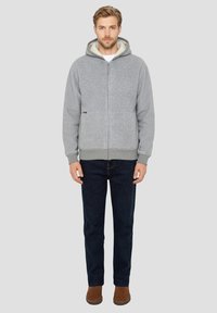 Grijze zip-up hoodie met gevoerde capuchon, geribbelde manchetten en zoom, met een klein logo op de linker voorkant. Gecombineerd met donkere jeans en bruine schoenen.