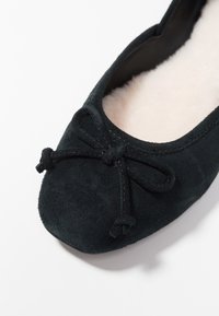 Černé semišové ploché boty s kulatou špičkou, ozdobené mašlí a měkkou vnitřní podšívkou. Textura se zdá být hladká a elegantní.