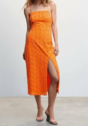 Kobieta w pomarańczowej sukience midi z wycięciami, z cienkimi ramiączkami i rozcięciem do uda, w połączeniu z przezroczystymi koturnami na szarej podłodze.