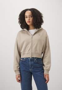 Sweat à capuche beige cropped avec fermeture éclair, doté d'une capuche et de poches frontales, associé à un jean bleu taille haute. Présente un petit logo de la marque sur la poitrine.