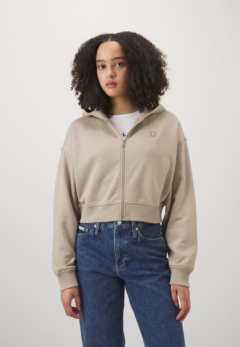 Sweat à capuche beige cropped avec fermeture éclair, doté d'une capuche et de poches frontales, associé à un jean bleu taille haute. Présente un petit logo de la marque sur la poitrine.