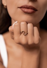 Goldene, wellenförmige Ring an einem Finger, mit glatter Oberfläche und polierter Finish, vor einem neutralen Hautton-Hintergrund.