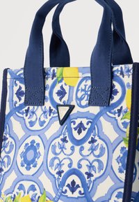 Τσάντα χειρός με ναυτικό μπλε λαβές, λευκό ύφασμα διακοσμημένο με μπ λε διακοσμητικά μοτίβα, κίτρινες λεμονιές και ένα μικρό μεταλλικό λογότυπο Guess.