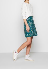 Spódnica w kwiatowy wzór w kolorze teal z dopasowaną talią, zestawiona z białą bluzką z długim rękawem i białymi trampkami za kostkę. Gładka tekstura materiału.