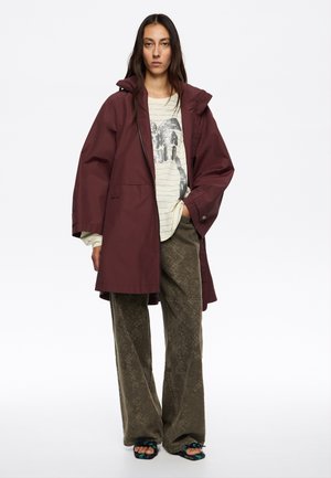 Chaqueta impermeable color burdeos con capucha, corte relajado, combinada con una camiseta gráfica de rayas y pantalones de pierna ancha texturizados en color verde oliva. Sandalias planas.