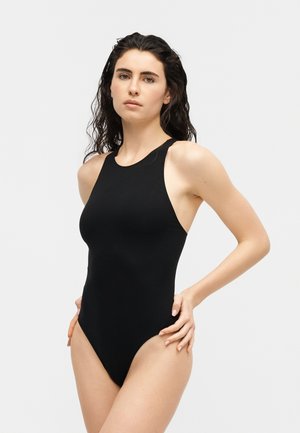 Νέος γυναίκα με μακριά σγουρά μαύρα μαλλιά που φοράει ένα μαύρο αμάνικο κορμάκι με ψηλό κόψιμο, στέκεται μπροστά σε απλό φωτεινό φόντο.